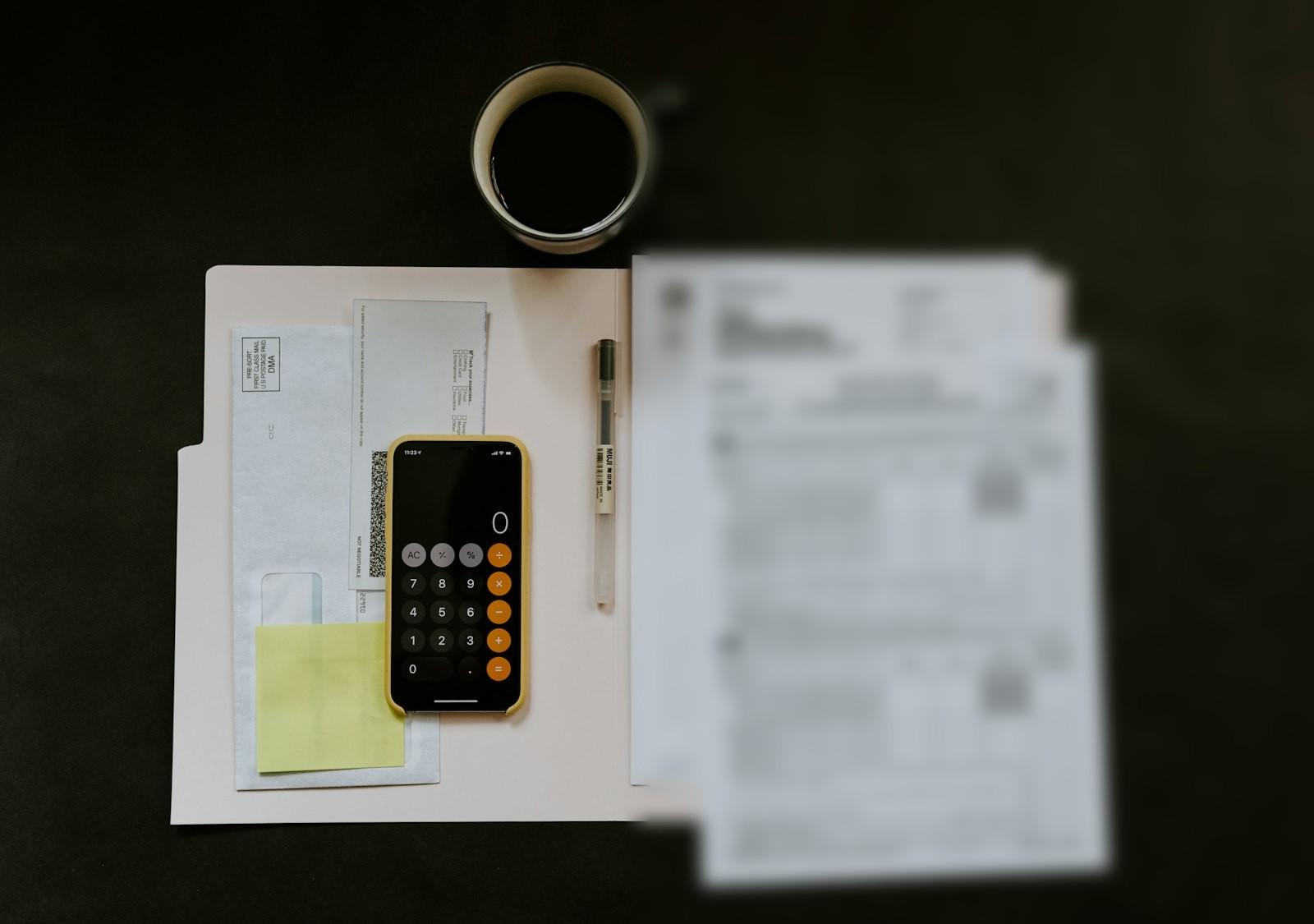 Pärm med kvitton och fakturor, en mobil med räknare och en kaffekopp på ett mörkt bord